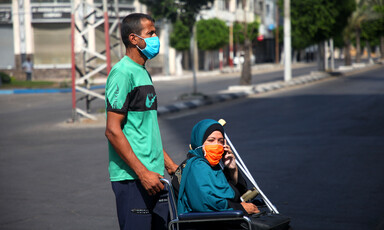 Man rolls woman on wheelchair, both wearing protective masks 