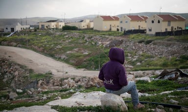 Youth sits with back turned to camera in front of Israeli settlement