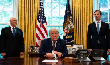 Man sits behind a desk, two men stand behind him 