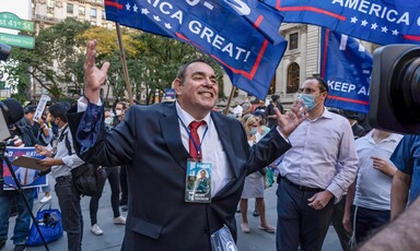 Man speaks with upraised arms with Trump flags behind him