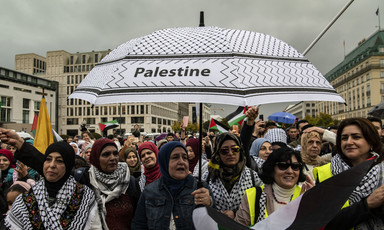 A group of women stand beneath a black-and-white umbrella emblazoned with the word 'Palestine'. 