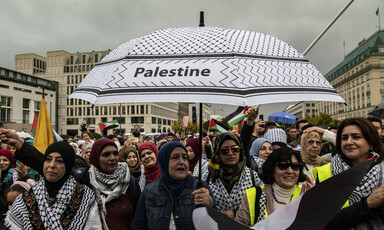 A group of women stand beneath a black-and-white umbrella emblazoned with the word 'Palestine'.