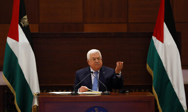 Mahmoud Abbas lifts hand in air while seated between two Palestinian flags