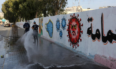 Men use hoses to spray sidewalk along wall with mural depicting a face in a coronavirus sphere and surgical masks 