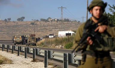 Armed Israeli soldier stands next to road with military vehicles behind him