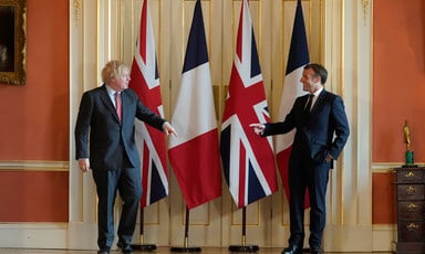 Boris Johnson and Emmanuel Macron stand in front of flags and point at each other 