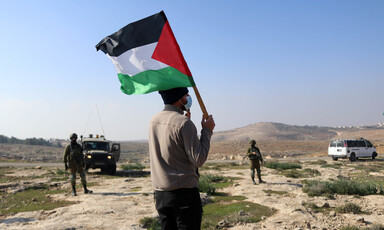 Man with back turned to camera holds Palestine flag while facing soldiers