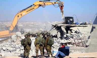 Heavily armed soldiers watch Hyundai bulldozer demolish a home while two men observe nearby 