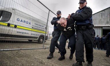 Police officers wearing baseball hats grab hold of a man by his arms 