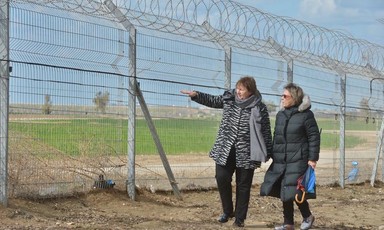 Two women stand beside large fence topped with barbed wire 