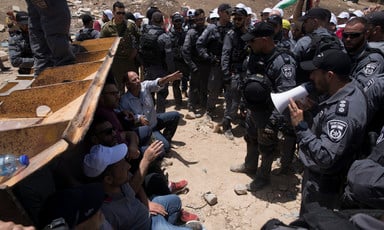 Armored police stand in line in front of people sitting in mouth of bulldozer