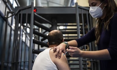 A standing, masked woman administers a vaccination to the shoulder of a man seated in front of metal turnstile