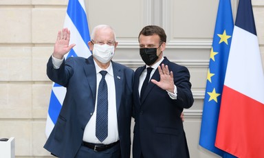 Reuven Rivlin and Emmanuel Macron, both wearing face masks, wave while standing in front of Israeli and EU flags