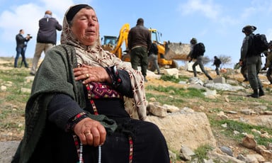 Woman sits on a rock with her hand on her chest as bulldozer operates in the background 