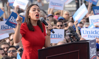 Woman speaks at podium in front of large crowd carrying signs 