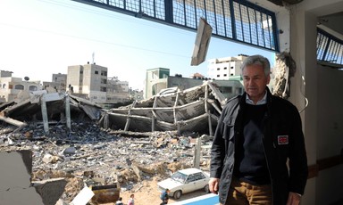 A man in a jacket stands with a ruined building in the background