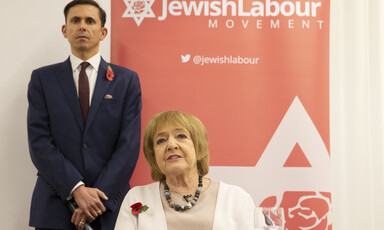 A man in a suit stands next to a woman sitting and talking