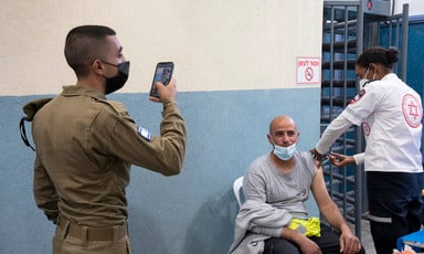 A uniformed soldier holds up a phone while a seated man receives a shot from a health care worker