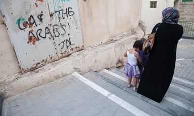 Woman and children walk past wall spray painted with graffiti reading Death to the Arabs!