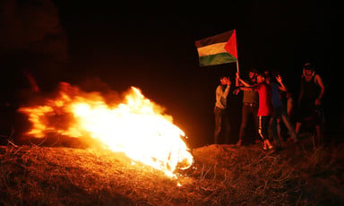 A small group of people stand next to a fire holding a Palestinian flag in the dark