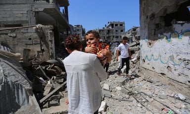 Girl holding baby is seen from back as she stands in bombed-out neighborhood