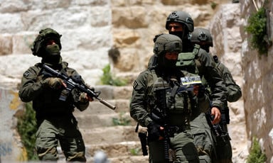 Four heavily armed Israeli soldiers stand near stone building 