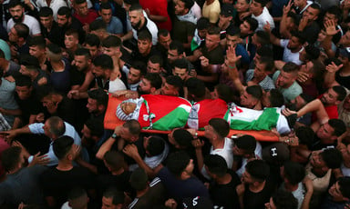 A large crowd of mourners carry a body wrapped in a Palestinian flag on a stretcher 