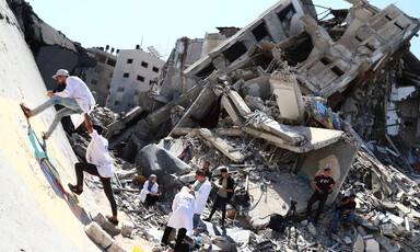 Two people climb part of a destroyed building, surrounded by other people