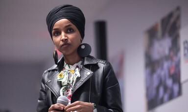 Ilhan Omar, wearing leather jacket, stands while holding microphone