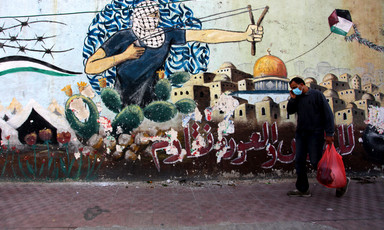 A man with a shopping bag walks past graffiti depicting the Dome of the Rock and keffiyeh-clad youth wielding a slingshot 
