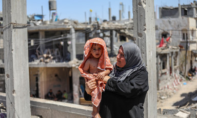Woman seen from waist up holds a toddler wrapped in a towel while standing in front of bombed-out buildings