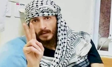 A man in a keffiyeh makes a V for victory sign with his hand