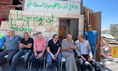 Seven men sit in front of a graffitied building, near rubble