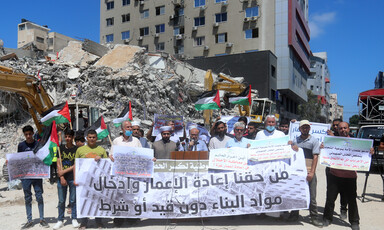 People hold banners and flags before a podium against a background of rubble 