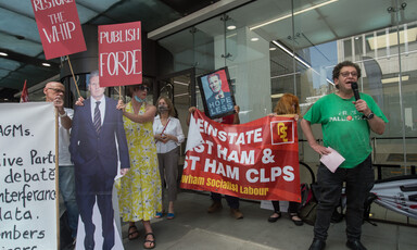 Man speaks into microphone as people around him hold signs and banners