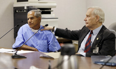 Man in suit places hand on shoulder of man in prison uniform seated next to him