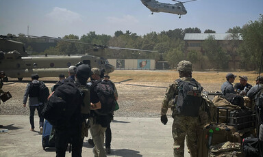 A helicopter lifts off as people holding baggage stand nearby