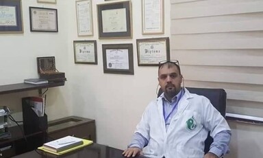 Man in medical uniform sits at desk with framed diplomas on wall behind him