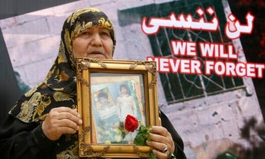 Woman holds framed photo of young man and rose in front of banner reading We will never forget in English and Arabic