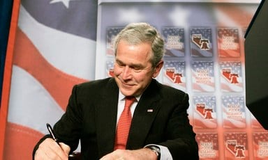 George W. Bush smirks as he signs document while sitting at table with American flag backdrop behind him