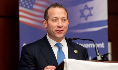 A man in a suit stands in front of US and Israeli flags