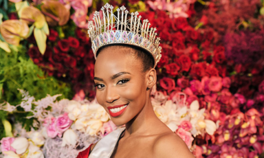 A woman wearing a crown and sash against a backdrop of flowers 