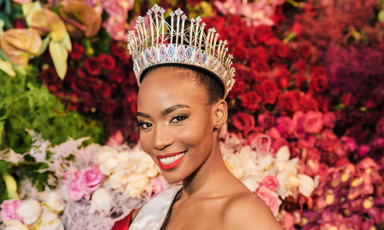 A woman wearing a crown and sash against a backdrop of flowers
