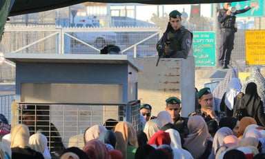 Soldier holding rifle stands behind elevated cinderblock above queue of Palestinians