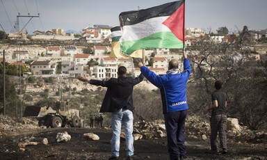 Two men hold up their arms while waving a Palestine flag and facing soldiers with settlement in background