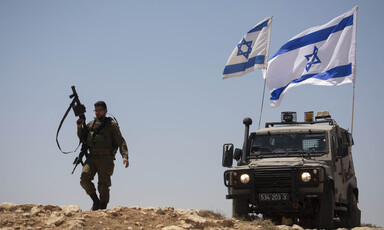 Soldier holds rifle in air while walking next to military Jeep flying two Israeli flags