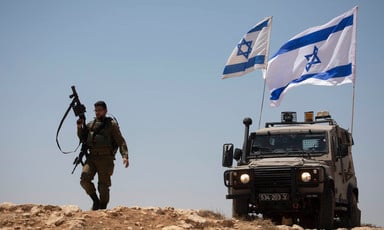 Soldier holds rifle in air while walking next to military Jeep flying two Israeli flags