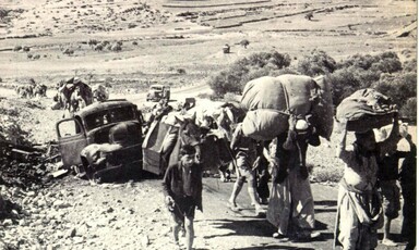 A black and white photo shows people on foot carrying heavy burdens passing a broken down vehicle