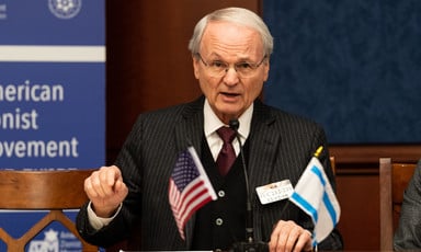 Photo of Morton Klein gesticulating with microphone and small American and Israeli flags in front of him