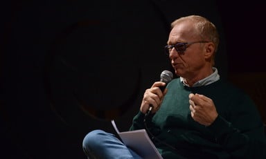 A man in glasses speaks into a microphone with a paper in his lap 